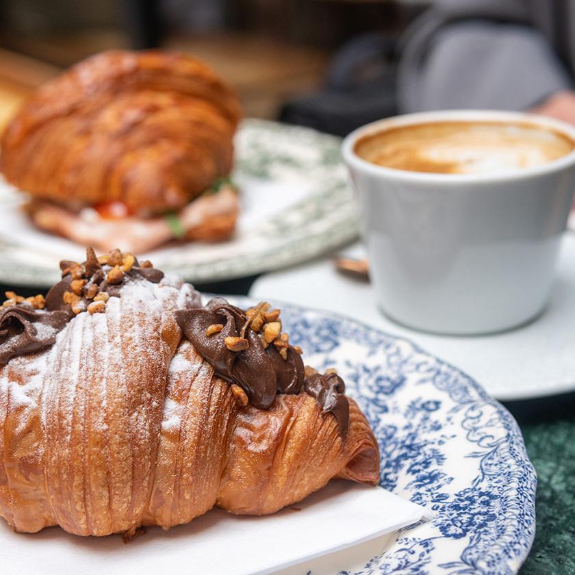 Croissant al cioccolato con granella, accompagnato da cappuccino su piatto decorato.